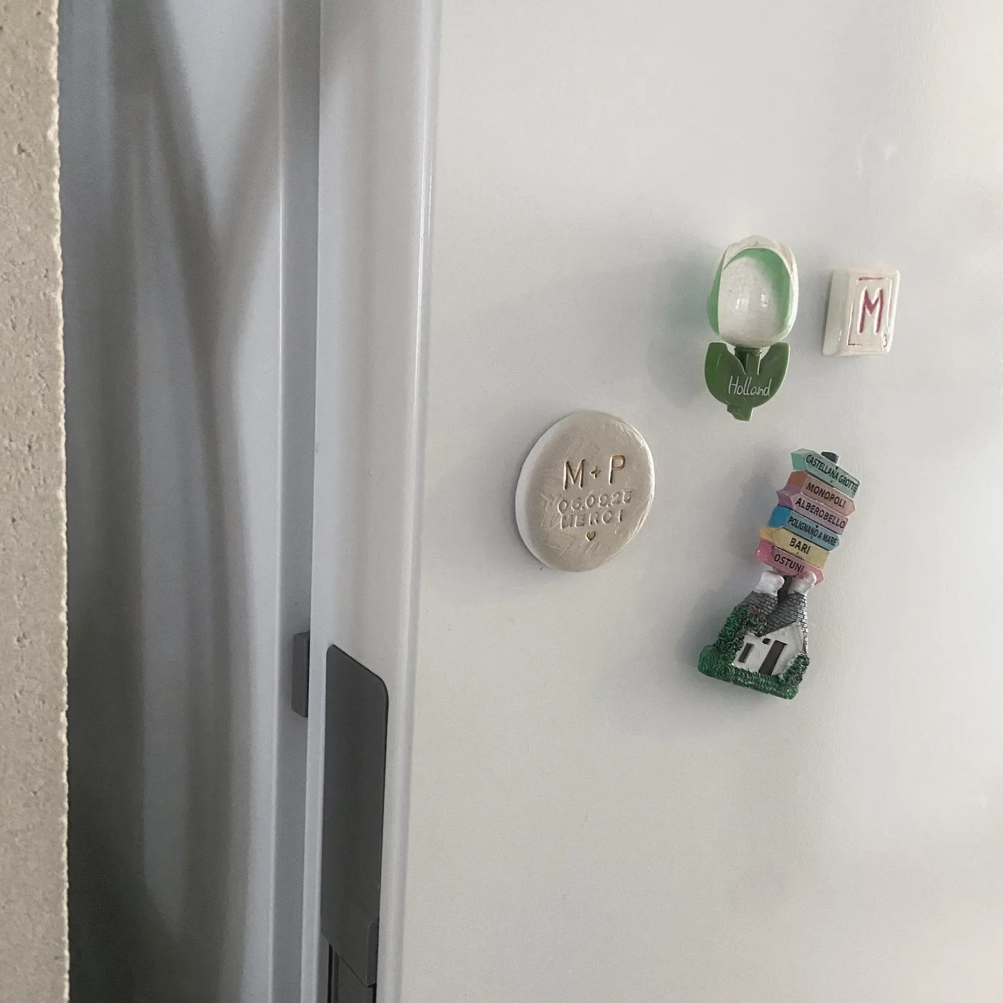Aimant en argile personnalisé posé sur un frigo, entouré d’autres magnets de voyage. Magnet fait main avec peinture dorée brillante pour effet céramique, idéal comme cadeau invité de mariage ou souvenir unique. Accessoire artisanal décoratif, élégant et personnalisé pour mariage, anniversaire ou baptême