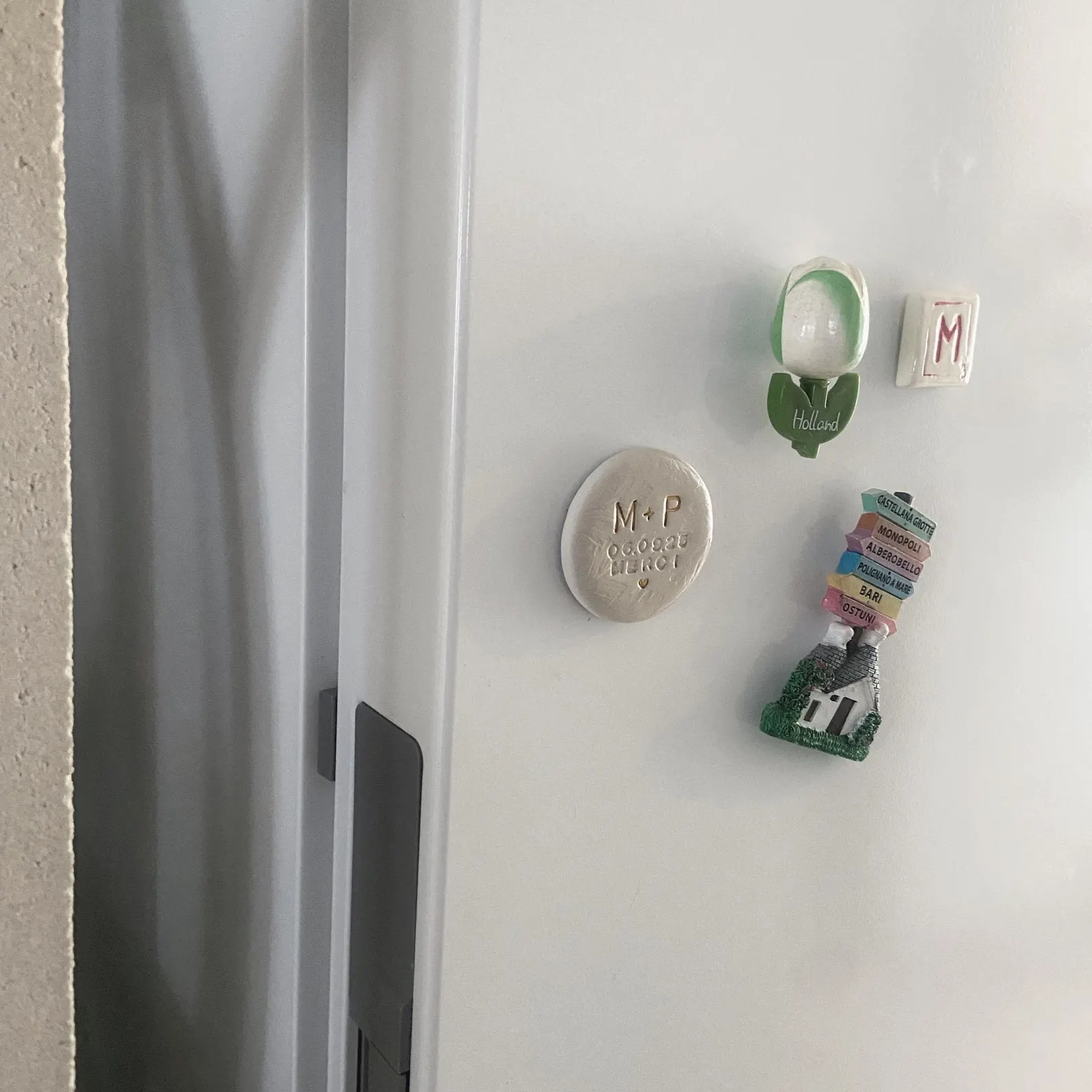 Aimant en argile personnalisé posé sur un frigo, entouré d’autres magnets de voyage. Magnet fait main avec peinture dorée brillante pour effet céramique, idéal comme cadeau invité de mariage ou souvenir unique. Accessoire artisanal décoratif, élégant et personnalisé pour mariage, anniversaire ou baptême