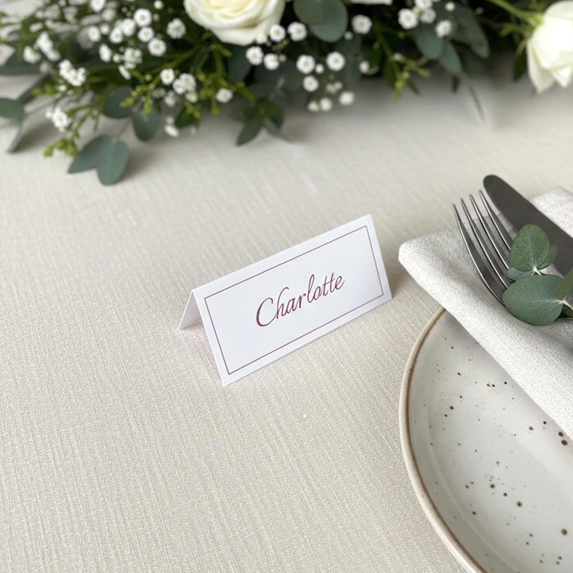 Marque-place de mariage personnalisé avec prénom 'Charlotte' en calligraphie bordeaux. Carte de table élégante avec bordure fine, posée sur une nappe en lin blanc à côté d'une assiette en céramique, d'un couvert et d'une décoration florale d'eucalyptus et de roses blanches