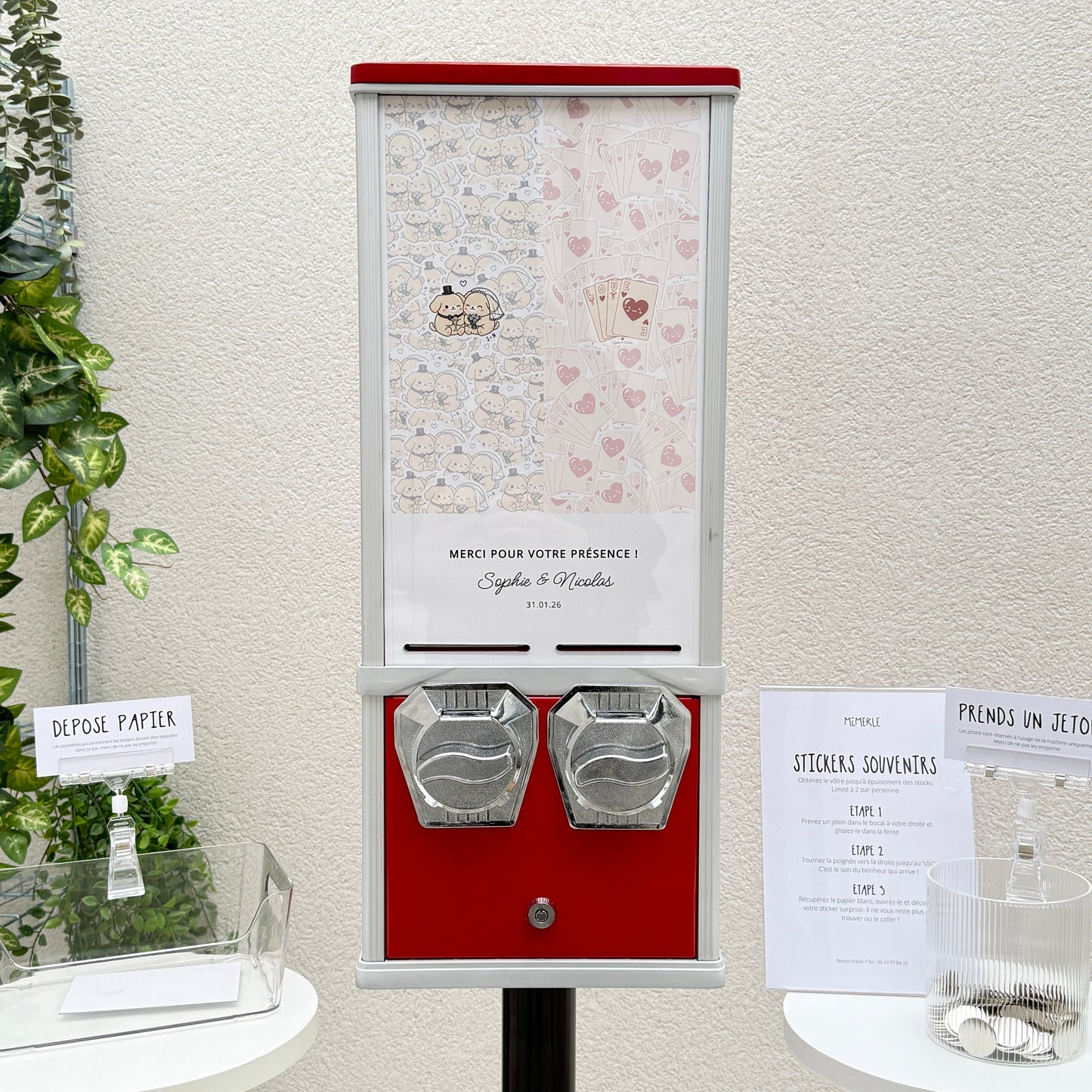 Un distributeur de stickers vintage rouge et gris personnalisé pour le mariage de Sophie et Nicolas, accompagné de deux petits guéridons avec des jetons et un bac pour le papier de protection.