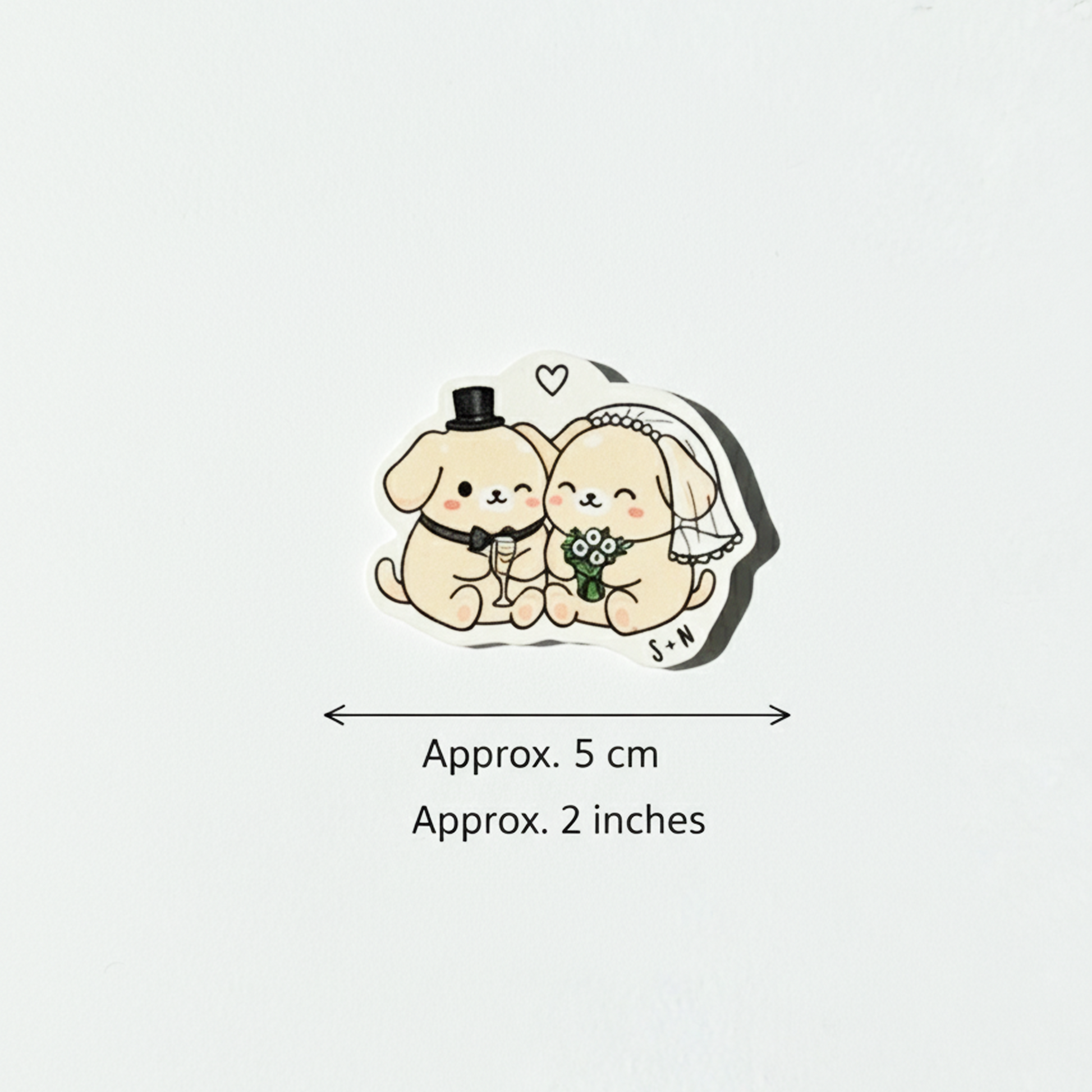 Un sticker illustrant un couple de chiots de couleur crème en tenue de mariage (un avec un chapeau haut de forme et un nœud papillon, l'autre avec un voile et un bouquet). Le sticker est posé sur un fond blanc épuré. En dessous, des flèches de mesure indiquent une largeur approximative de 5 cm ou 2 pouces.