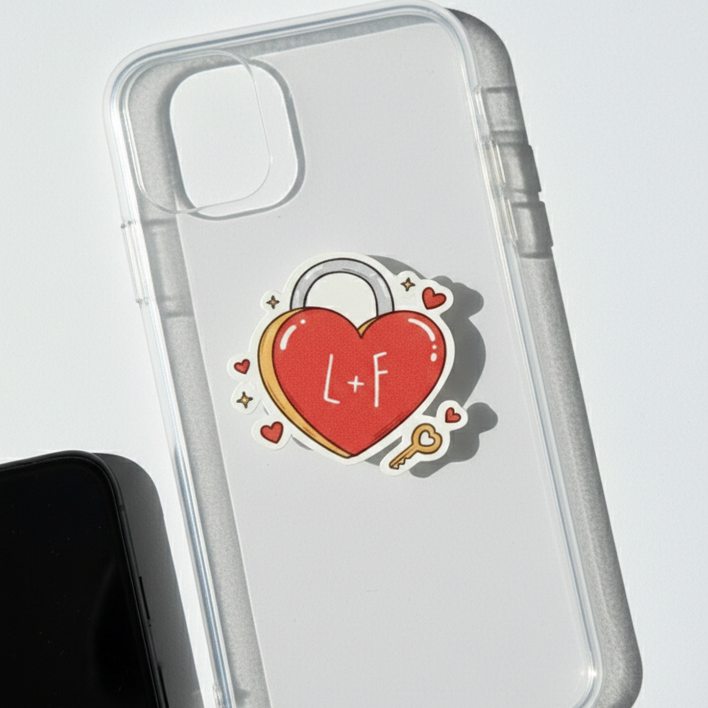 Une photo en vue de dessus montrant un smartphone noir inséré dans une coque transparente. Au centre de la coque est collé un sticker en forme de cadenas en cœur rouge, sur lequel sont inscrites les lettres 'L+F' en blanc. Le sticker est entouré de petits motifs de cœurs et d'étincelles, avec une petite clé dorée sur le côté. L'ensemble repose sur une surface blanche épurée
