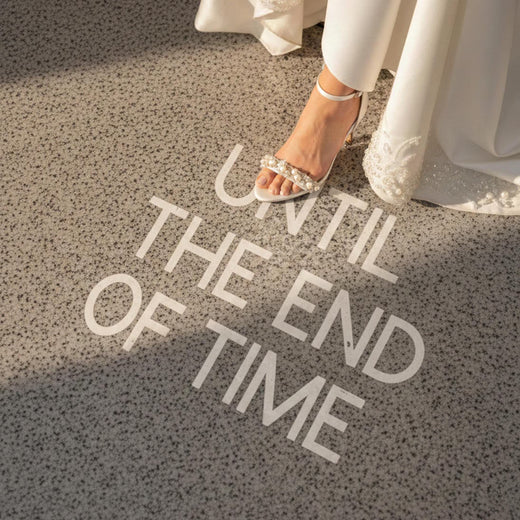 Vue de dessus d'une mariée portant des sandales argentées minimalistes, posant son pied sur un sol en granite élégant où est inscrit le message romantique "UNTIL THE END OF TIME" en lettres blanches. Ambiance de mariage chic et intemporelle. — Memerle, papeterie artisanale personnalisée (Paris, France & international)